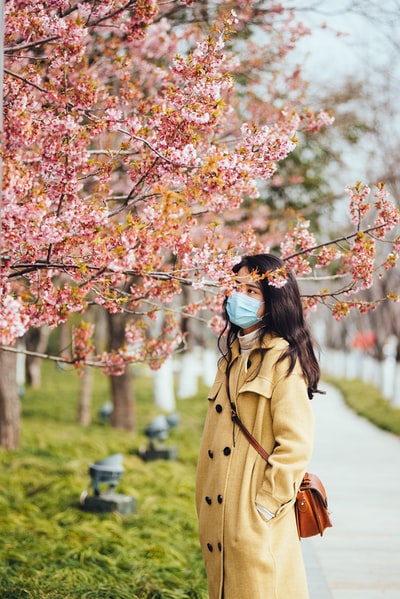 白天站在粉红色樱花树下的棕色外套女人